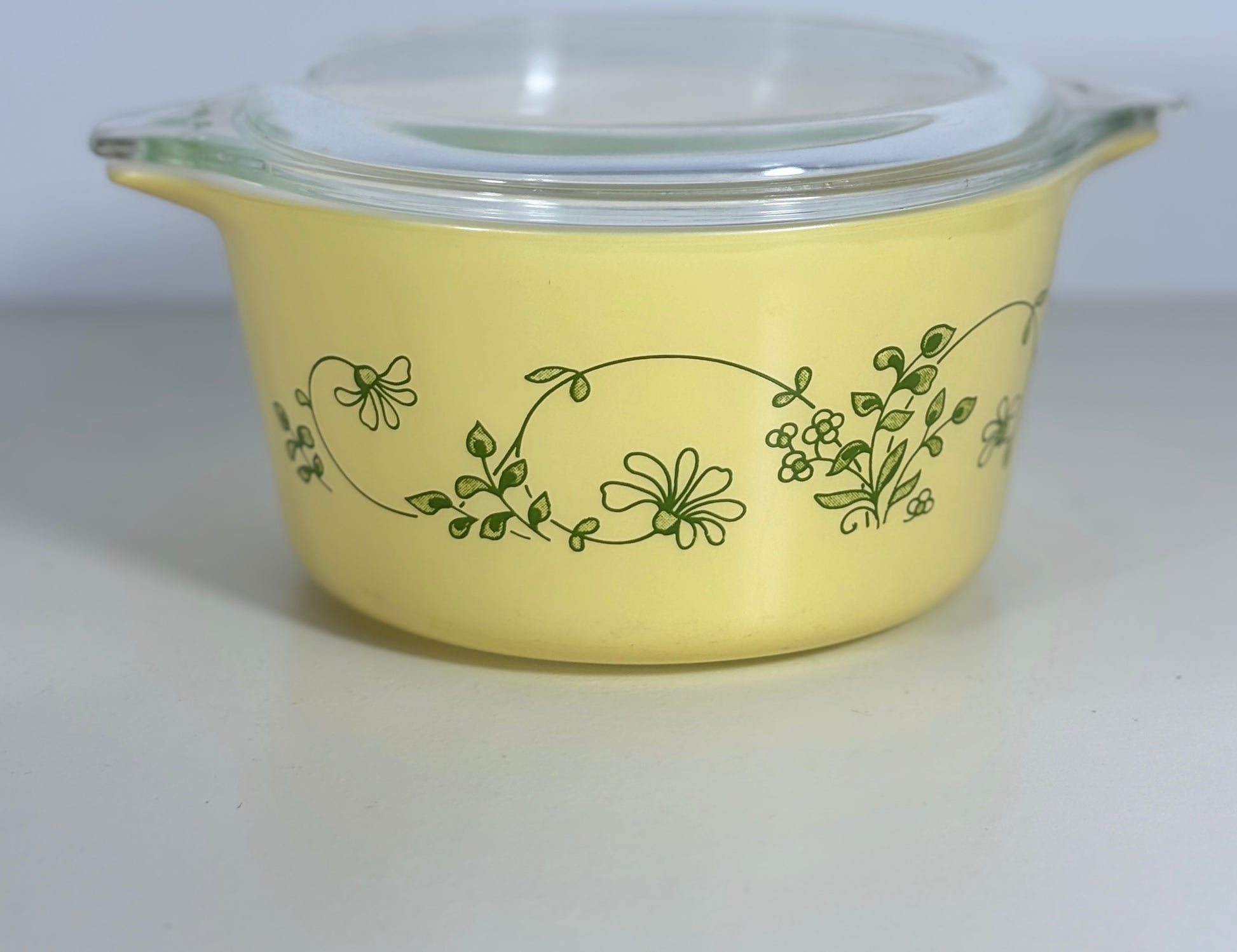 A yellow glass casserole dish with a clear glass lid, decorated with a green floral vine pattern, set against a plain light gray background.