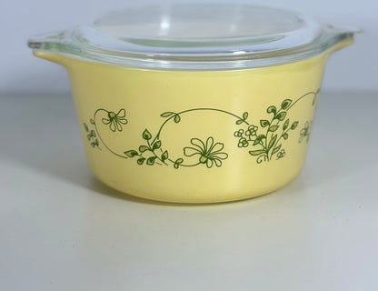 A yellow glass casserole dish with a clear glass lid, decorated with a green floral vine pattern, set against a plain light gray background.