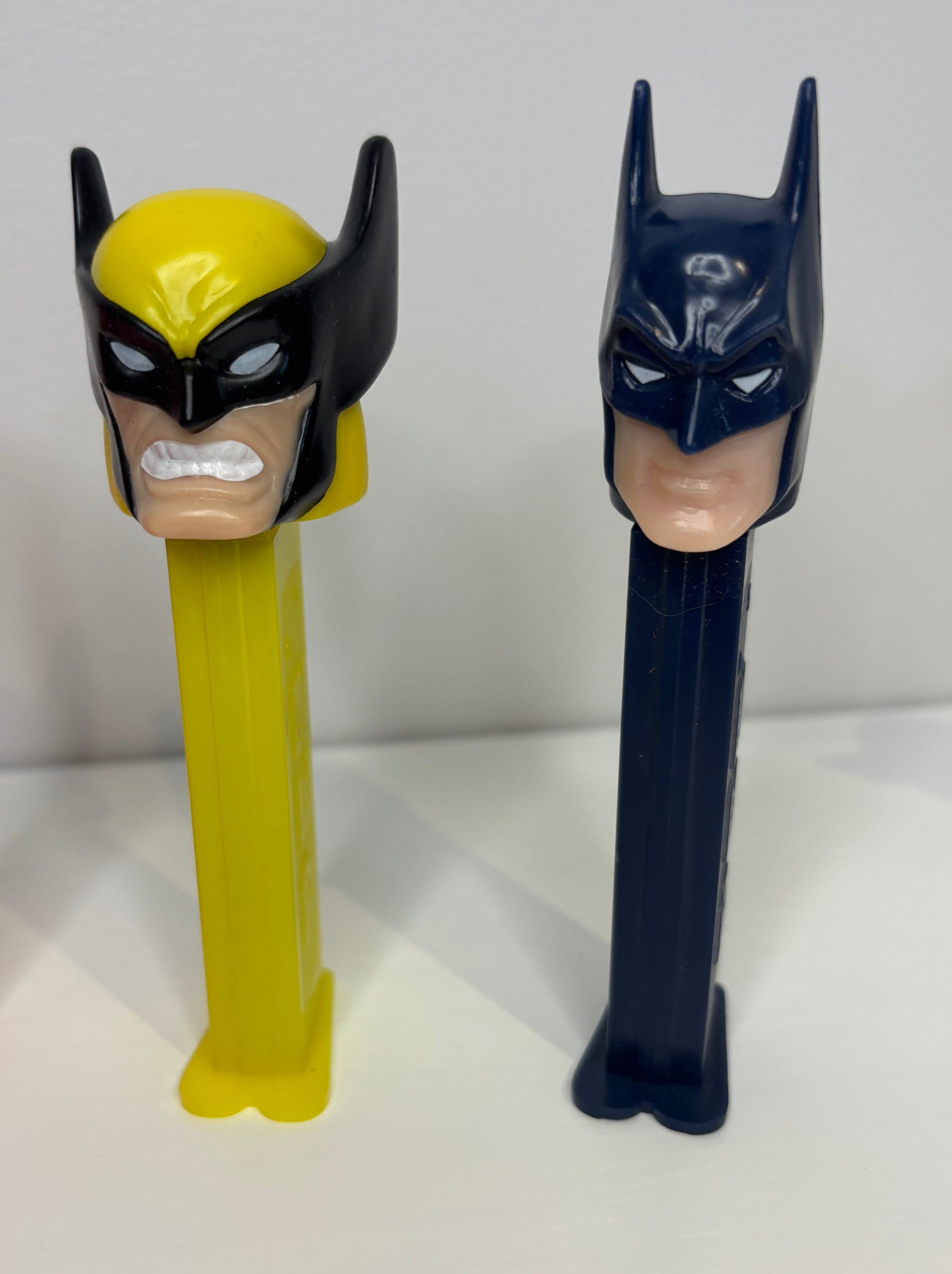 Two superhero-themed PEZ dispensers, one yellow with black mask and one blue with black mask, on a white background.