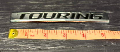 Metallic 'TOURING' emblem next to a ruler on a dark surface