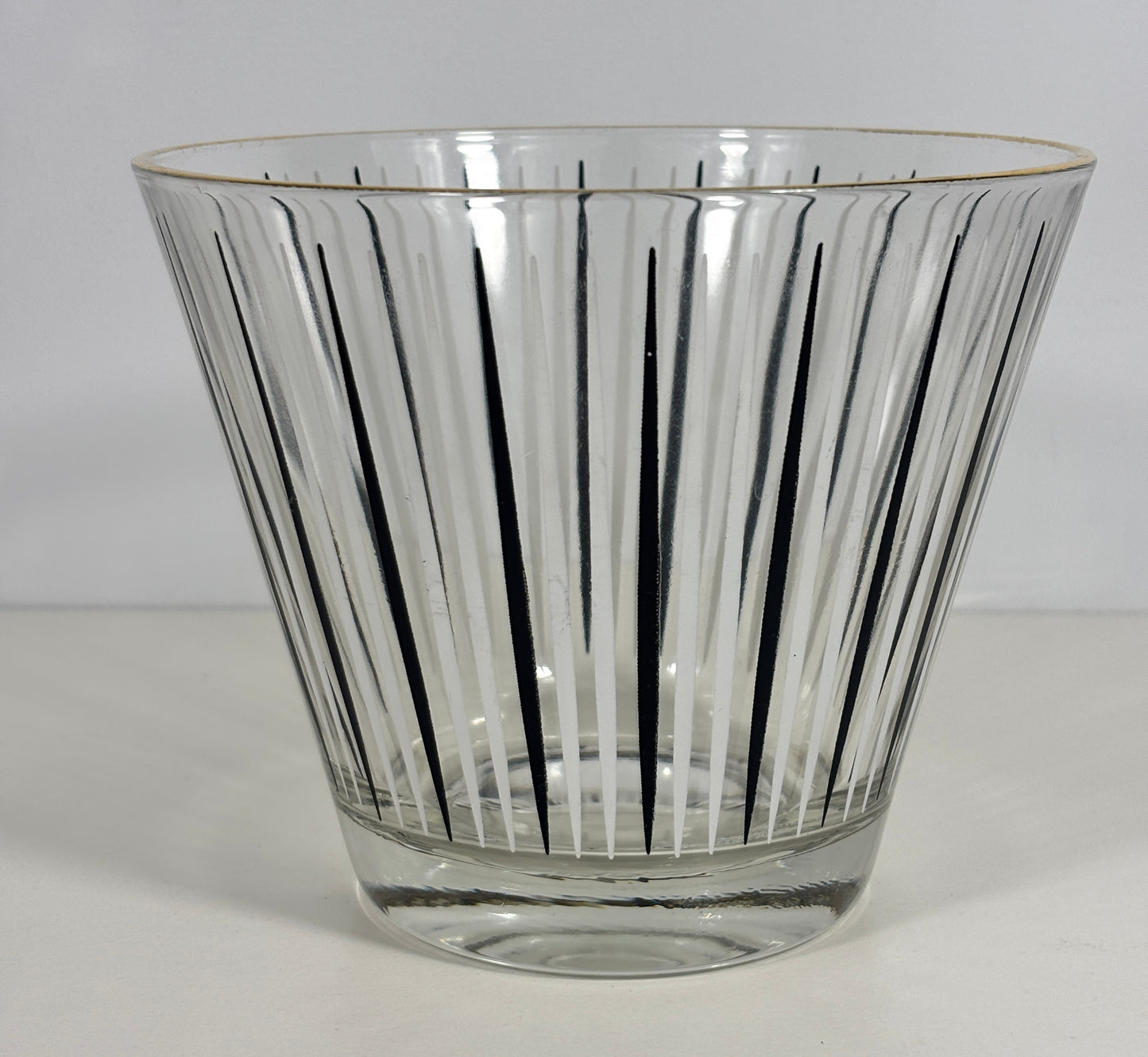 A clear glass ice bucket with a flared rim, featuring vertical black radial lines and a thin gold rim accent, set against a plain light gray background.