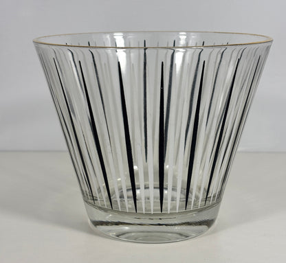 A clear glass ice bucket with a flared rim, featuring vertical black radial lines and a thin gold rim accent, set against a plain light gray background.
