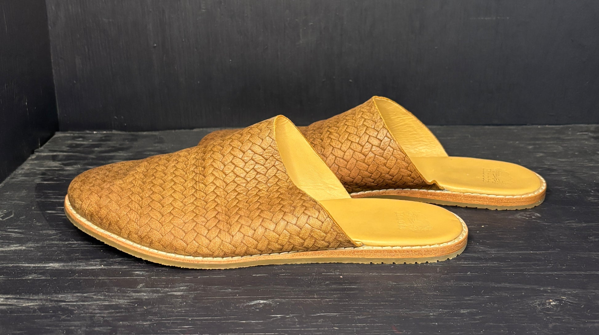 Pair of brown woven mules on a dark wooden surface with a black background