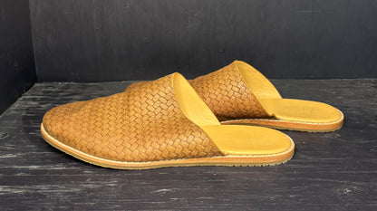 Pair of brown woven mules on a dark wooden surface with a black background