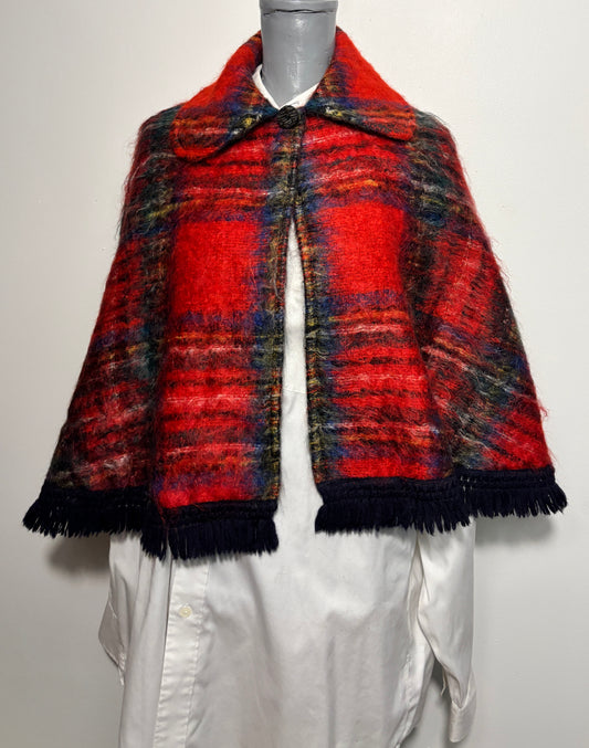 A red and navy plaid fringed cape sweater displayed on a gray mannequin torso wearing a white shirt. The cape features a thick textured wool-like fabric, a collar, and a button closure.