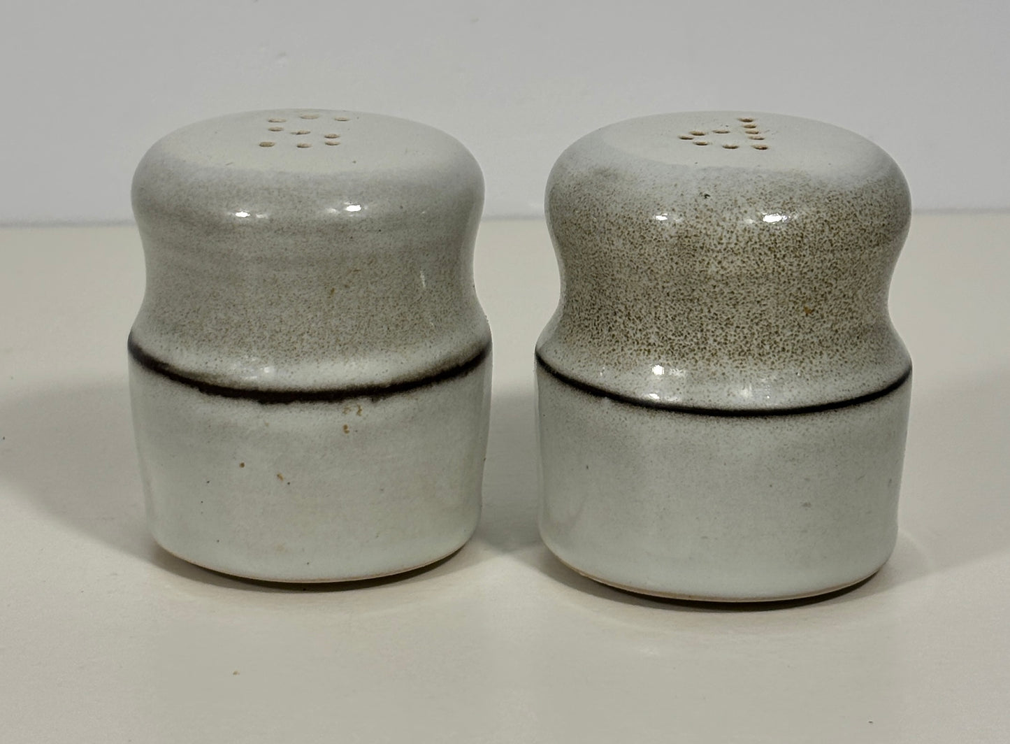 A pair of ceramic shaker salt and pepper mills with a speckled light gray and white glaze, featuring a dark brown band around the middle and a small perforation on top.