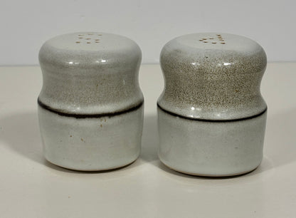 A pair of ceramic shaker salt and pepper mills with a speckled light gray and white glaze, featuring a dark brown band around the middle and a small perforation on top.