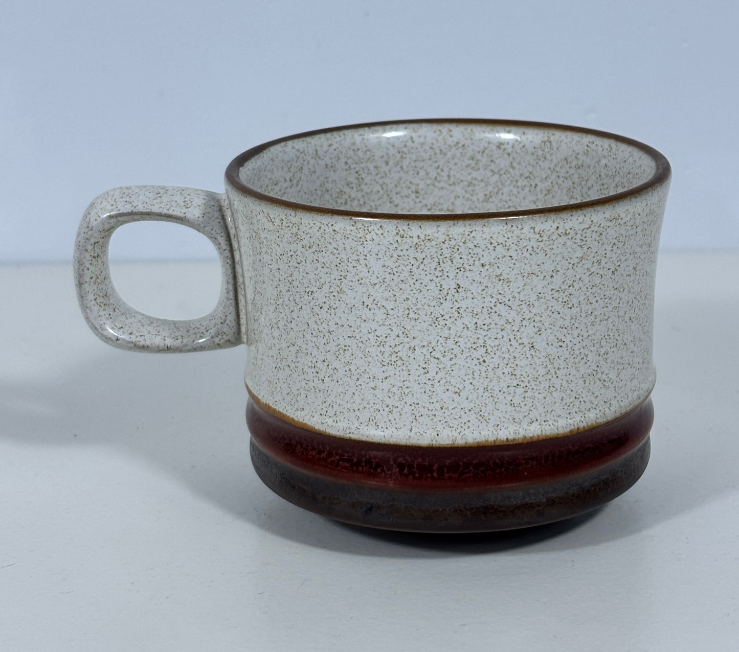 A ceramic mug with a speckled off-white glaze, a dark brown rim, and a thick dark brown band around the lower half, set against a plain light gray background.
