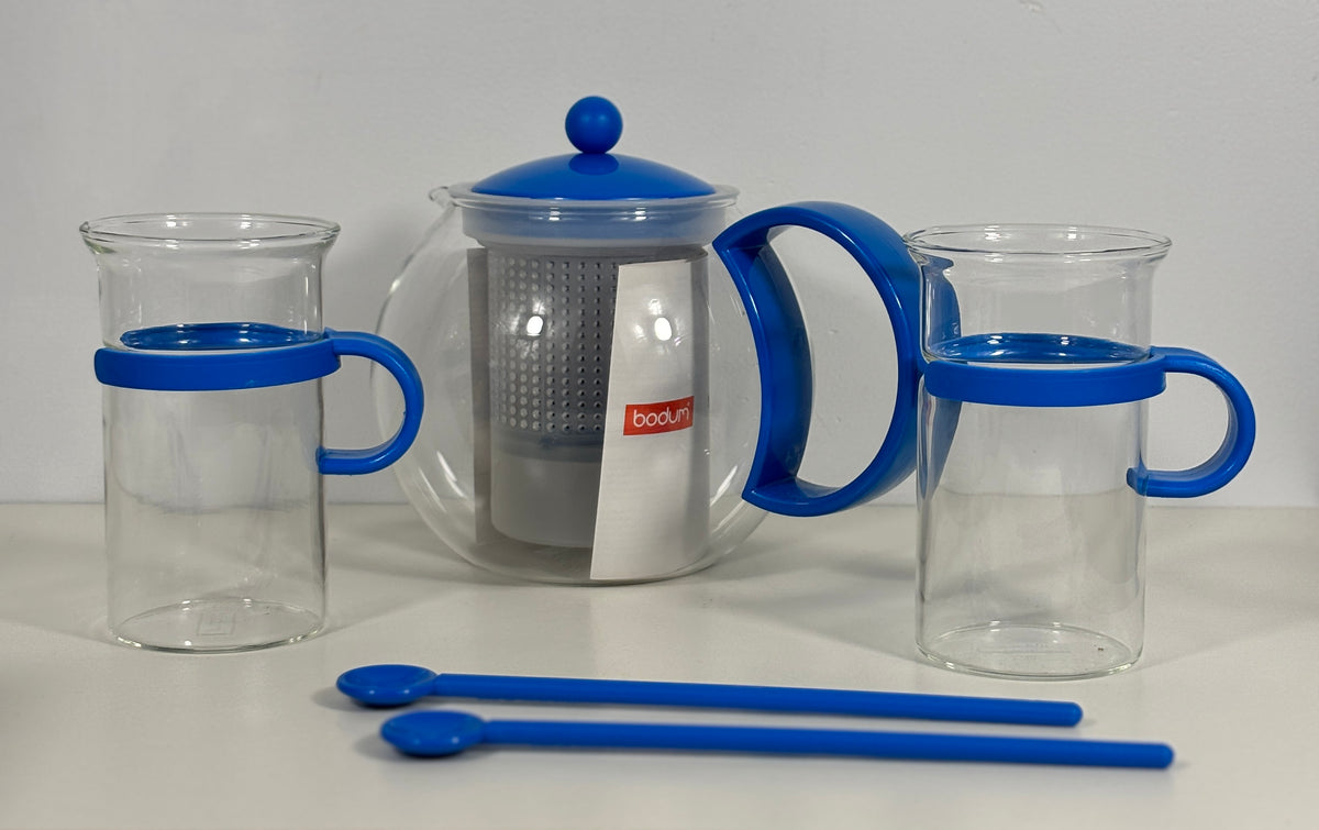 A glass tea set with blue plastic handles and lids, including a teapot with a metal infuser, two clear glass mugs with blue handles, and two blue spoons, displayed on a white surface against a light gray background.
