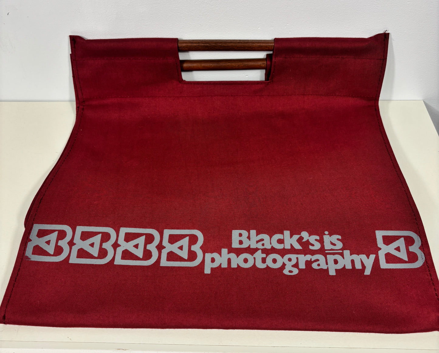 A dark red fabric tote bag with a double wooden handle, featuring the text 'Black's is photography' printed in gray on the front.