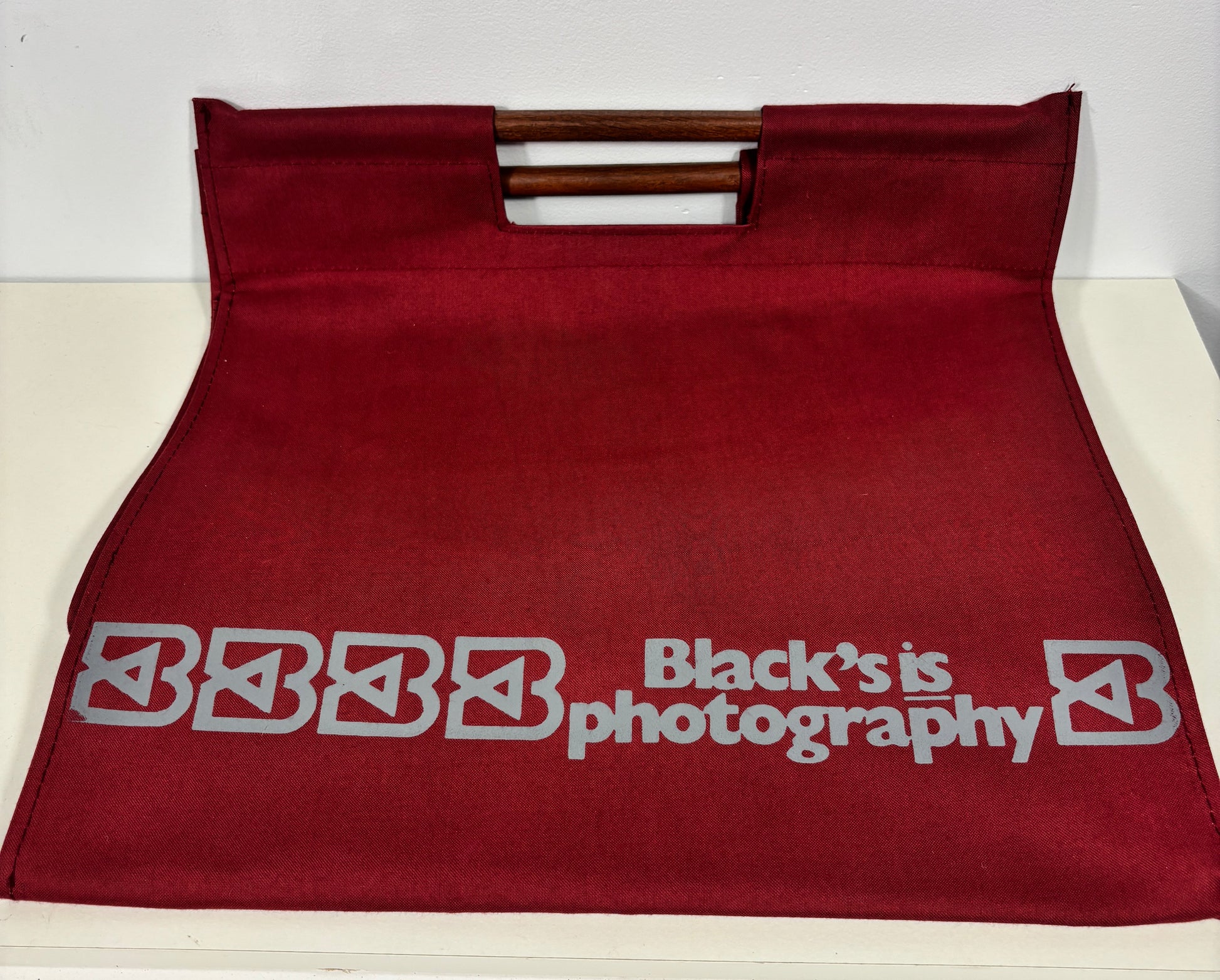 A dark red fabric tote bag with a double wooden handle, featuring the text 'Black's is photography' printed in gray on the front.