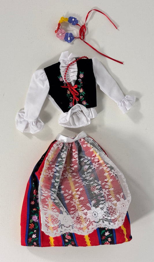 A traditional doll or costume outfit laid flat on a light background, featuring a black embroidered vest with red trim, a white long-sleeve blouse with ruffles, a multicolored patterned skirt with lace overlay, and a small floral hair accessory with red ties.