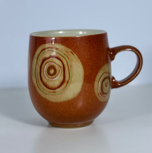 A brown ceramic mug with a glossy finish, decorated with two concentric circular wood-grain style patterns in a light beige color, set against a plain light gray background.