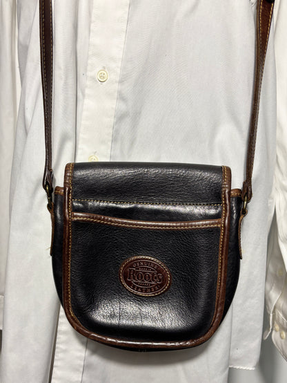 A dark brown leather crossbody bag with a prominent brown oval logo patch on the front pocket, worn over the chest of a person wearing a white button-down shirt.