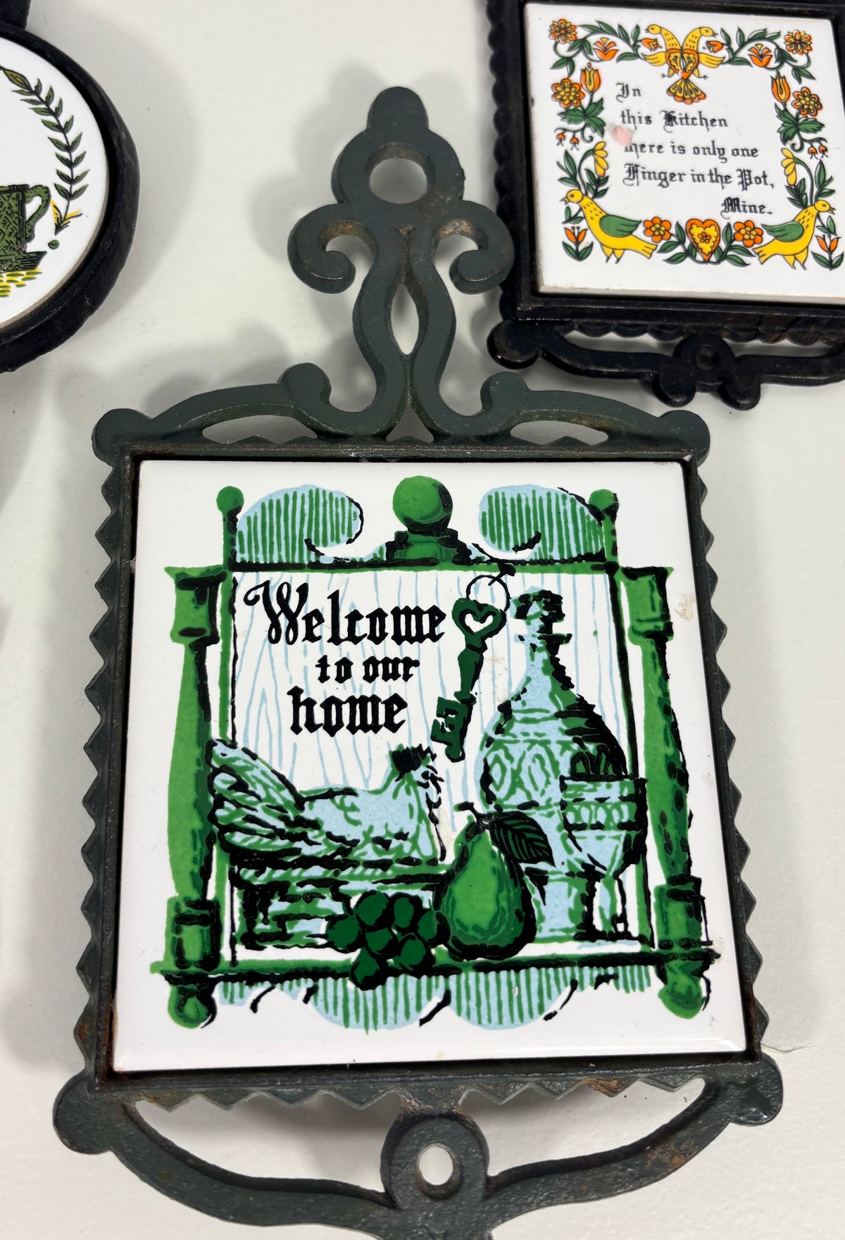 A decorative square wall plaque with an ornate dark metal frame and a white center panel featuring green illustrated artwork and the words 'Welcome to our home.' Part of another framed decorative piece is visible at the top right, and a cropped portion of another plaque appears at the left edge.
