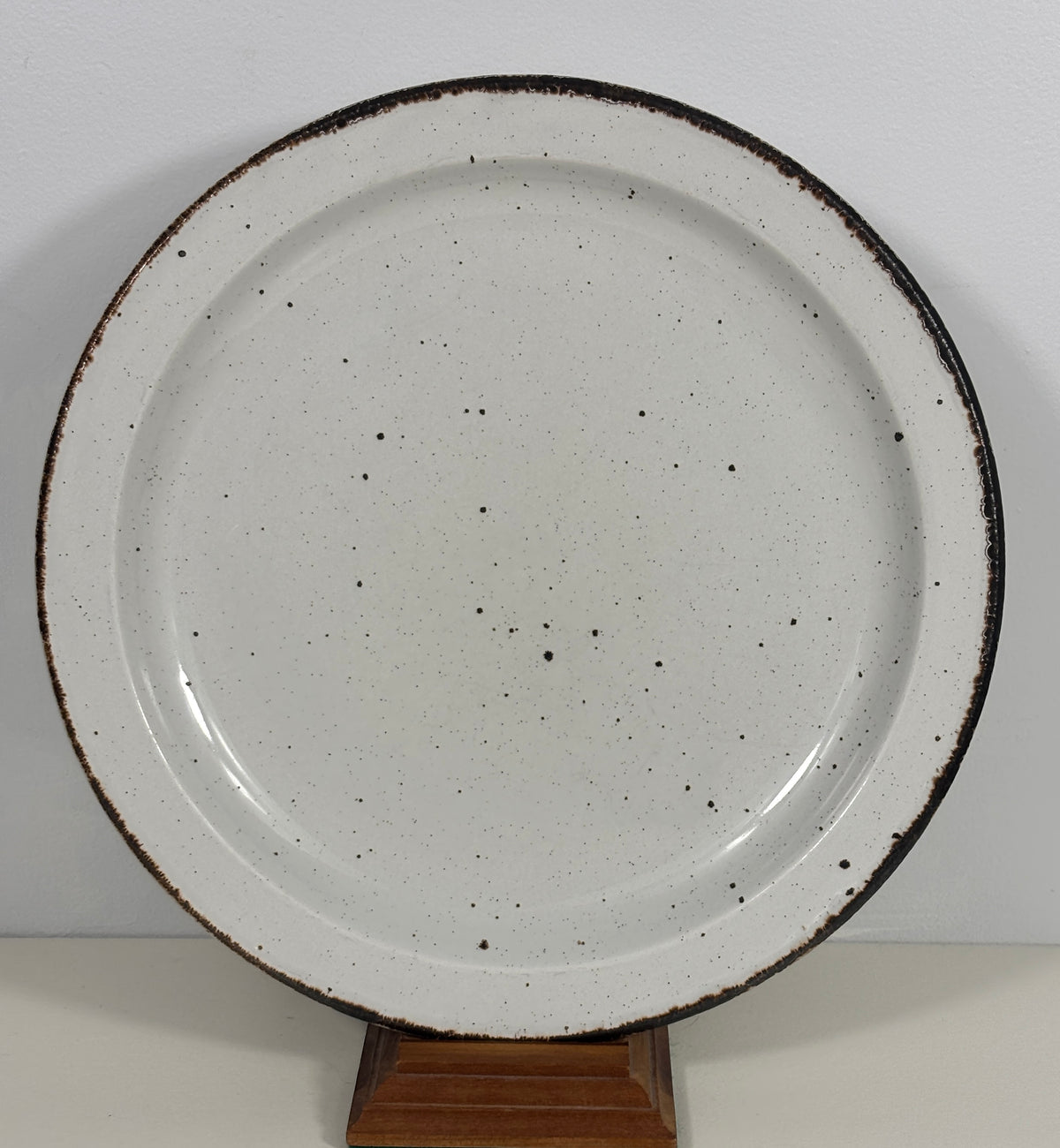 A round ceramic plate with a speckled off-white glaze and a rustic dark brown rim, displayed on a small wooden stand against a plain light background.