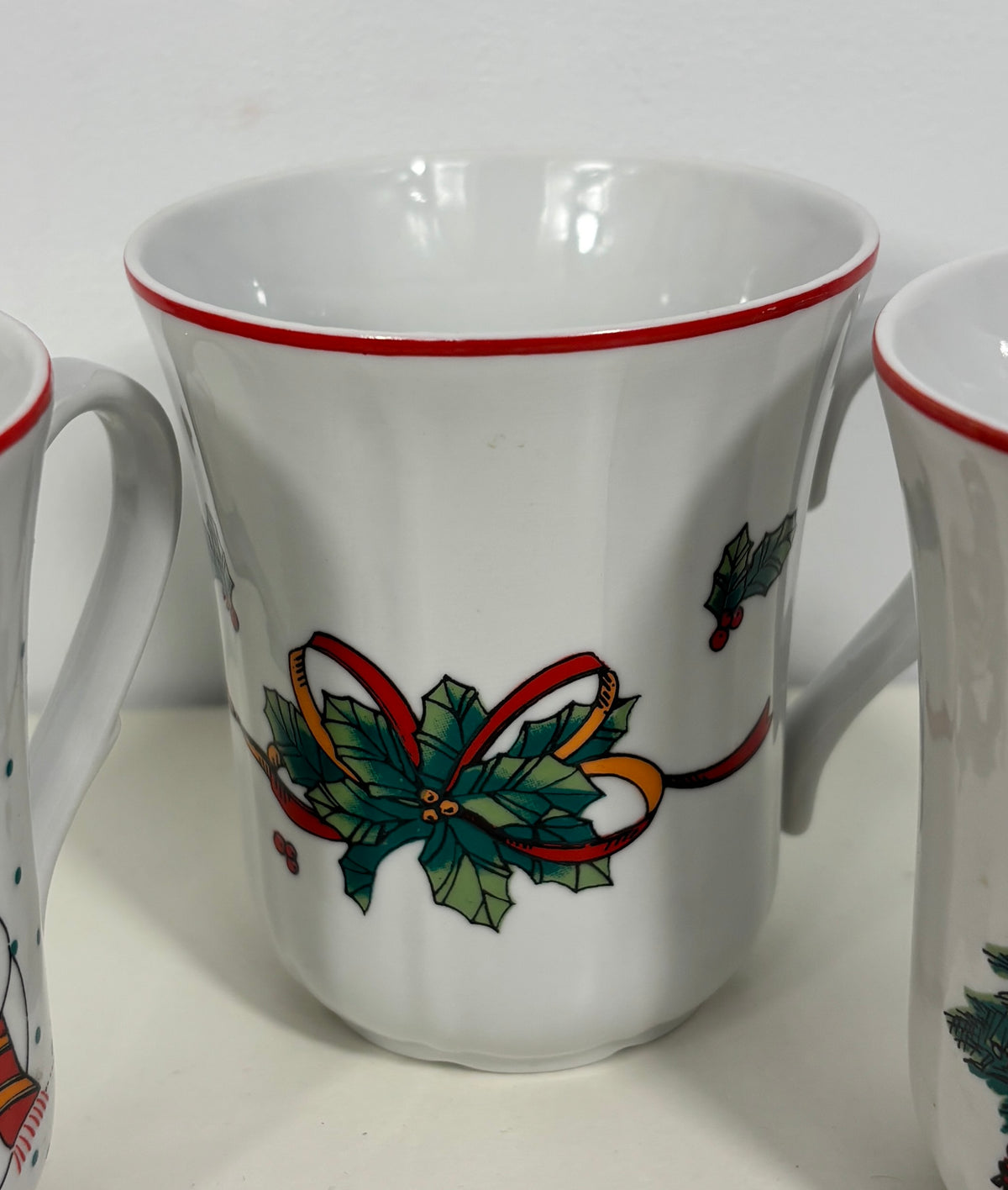 A white ceramic mug with a red rim and a green holly and red ribbon holiday design on the front, shown on a white surface with other mugs partially visible on both sides.