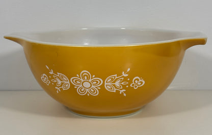 A vintage yellow glass mixing bowl with a white interior and a white floral pattern on the exterior, set against a plain light gray background.