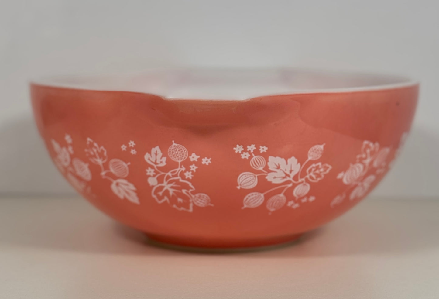 A coral orange glass bowl with a white floral and berry pattern, photographed against a neutral light gray background.