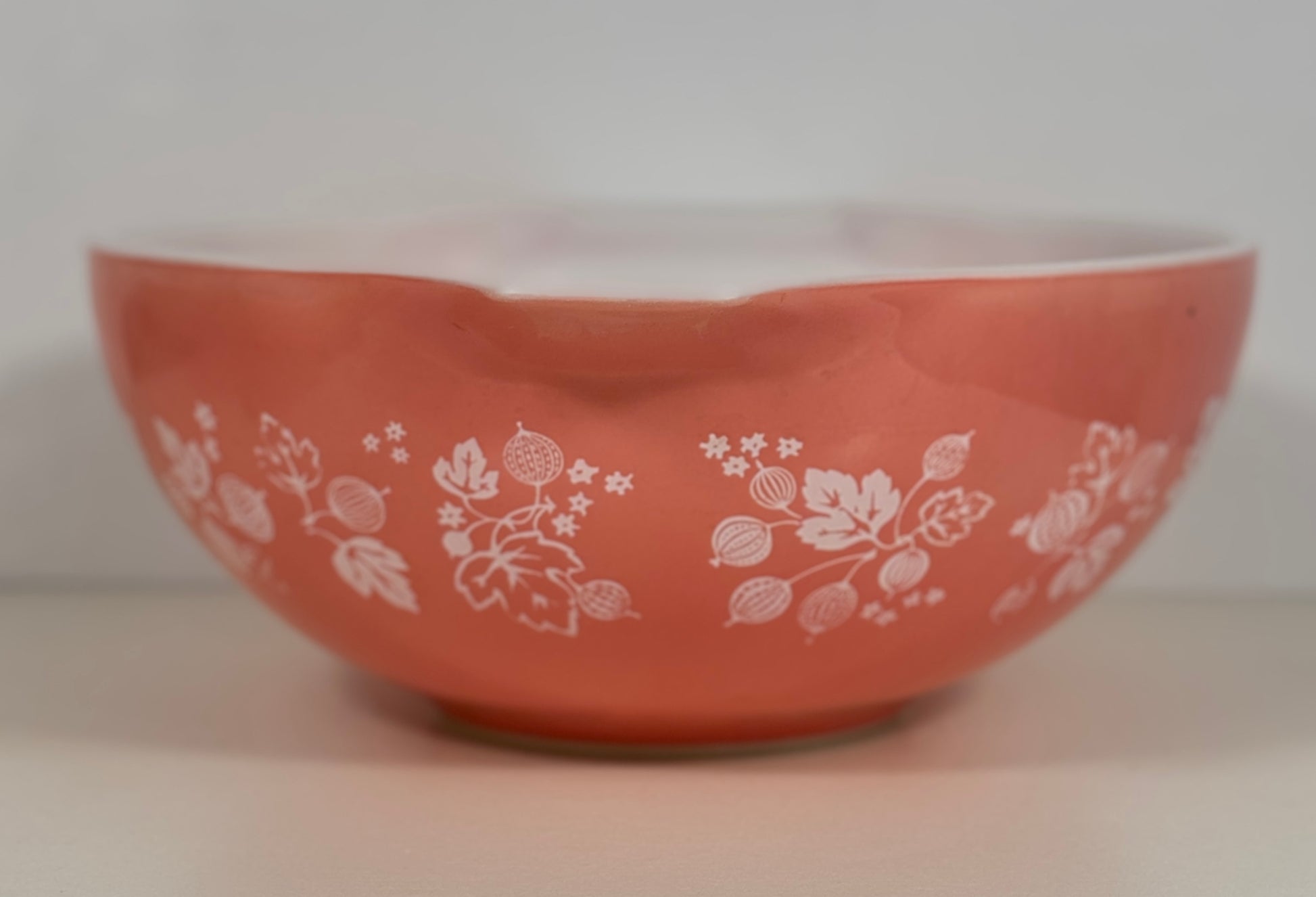 A coral orange glass bowl with a white floral and berry pattern, photographed against a neutral light gray background.