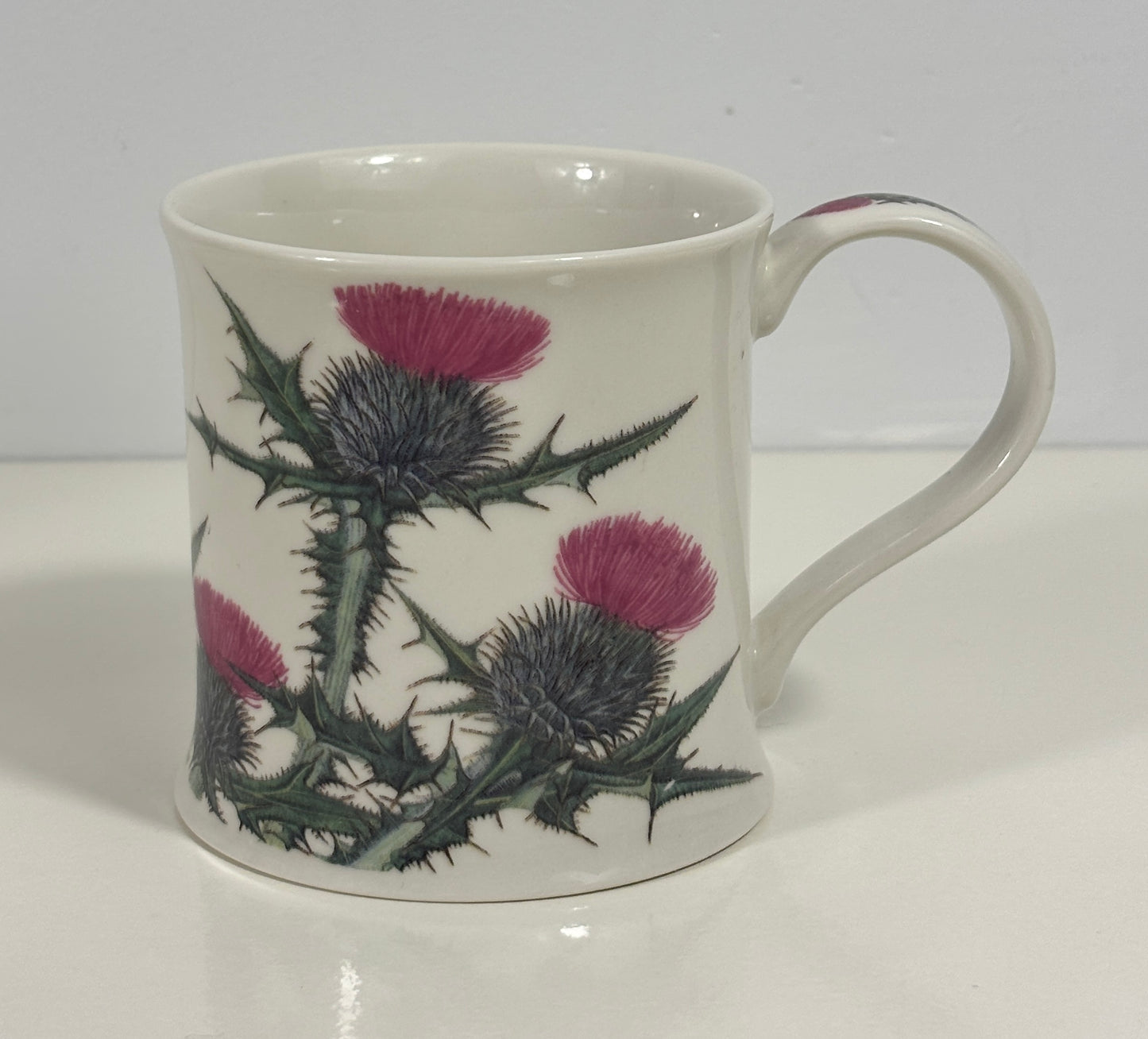 A white ceramic mug decorated with a botanical pattern of purple thistles and green thistle stems, set against a plain white background.