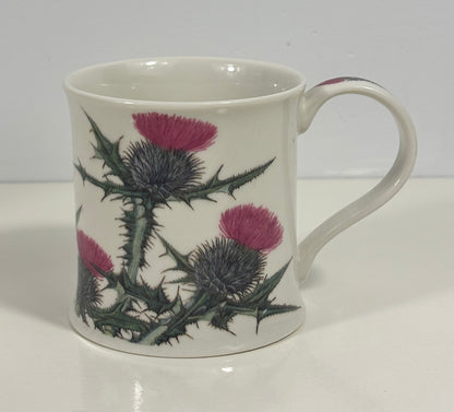 A white ceramic mug decorated with a botanical pattern of purple thistles and green thistle stems, set against a plain white background.