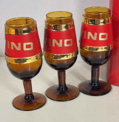 Three amber-brown stemmed glasses with red bands and gold trim, each printed with the word 'VINO' in gold, displayed on a light surface.