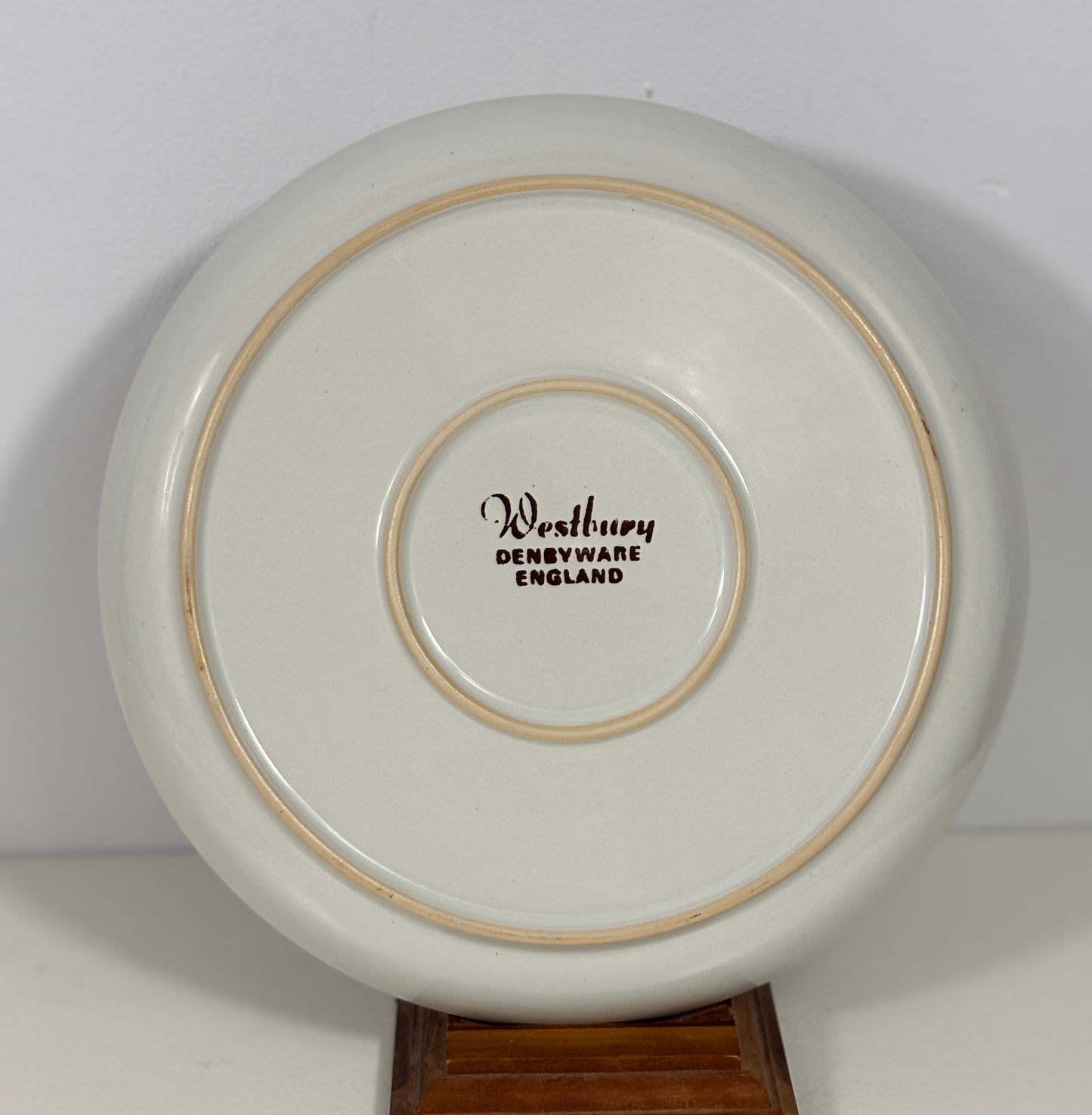The underside of a round ceramic plate or dish, showing a glossy off-white glaze with two thin tan rings and the printed text 'Westbury DENBYWARE ENGLAND' in the center, displayed on a wooden stand against a plain light background.
