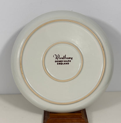 The underside of a round ceramic plate or dish, showing a glossy off-white glaze with two thin tan rings and the printed text 'Westbury DENBYWARE ENGLAND' in the center, displayed on a wooden stand against a plain light background.