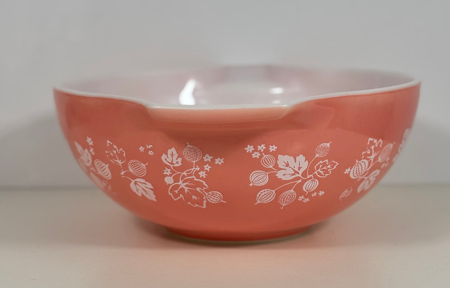 A coral pink Pyrex mixing bowl with a white floral pattern, photographed against a neutral light gray background.