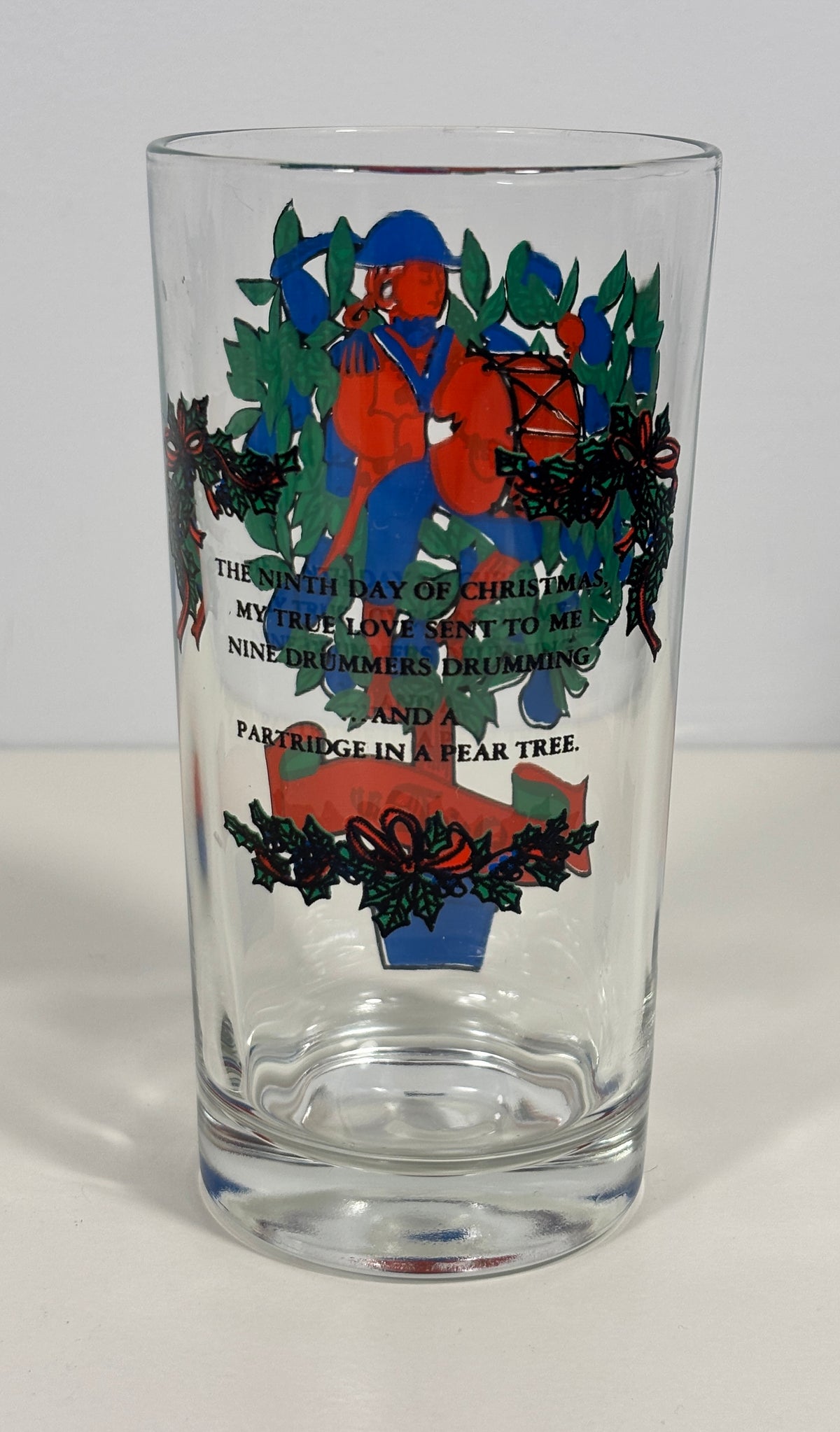 A clear glass drinking tumbler with a Christmas-themed printed design featuring a figure in red and blue, green holly leaves, and black text on a white background.
