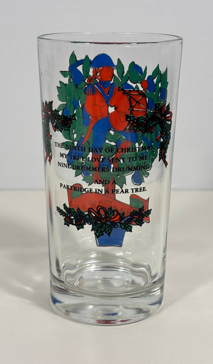 A clear glass drinking tumbler with a Christmas-themed printed design featuring a figure in red and blue, green holly leaves, and black text on a white background.