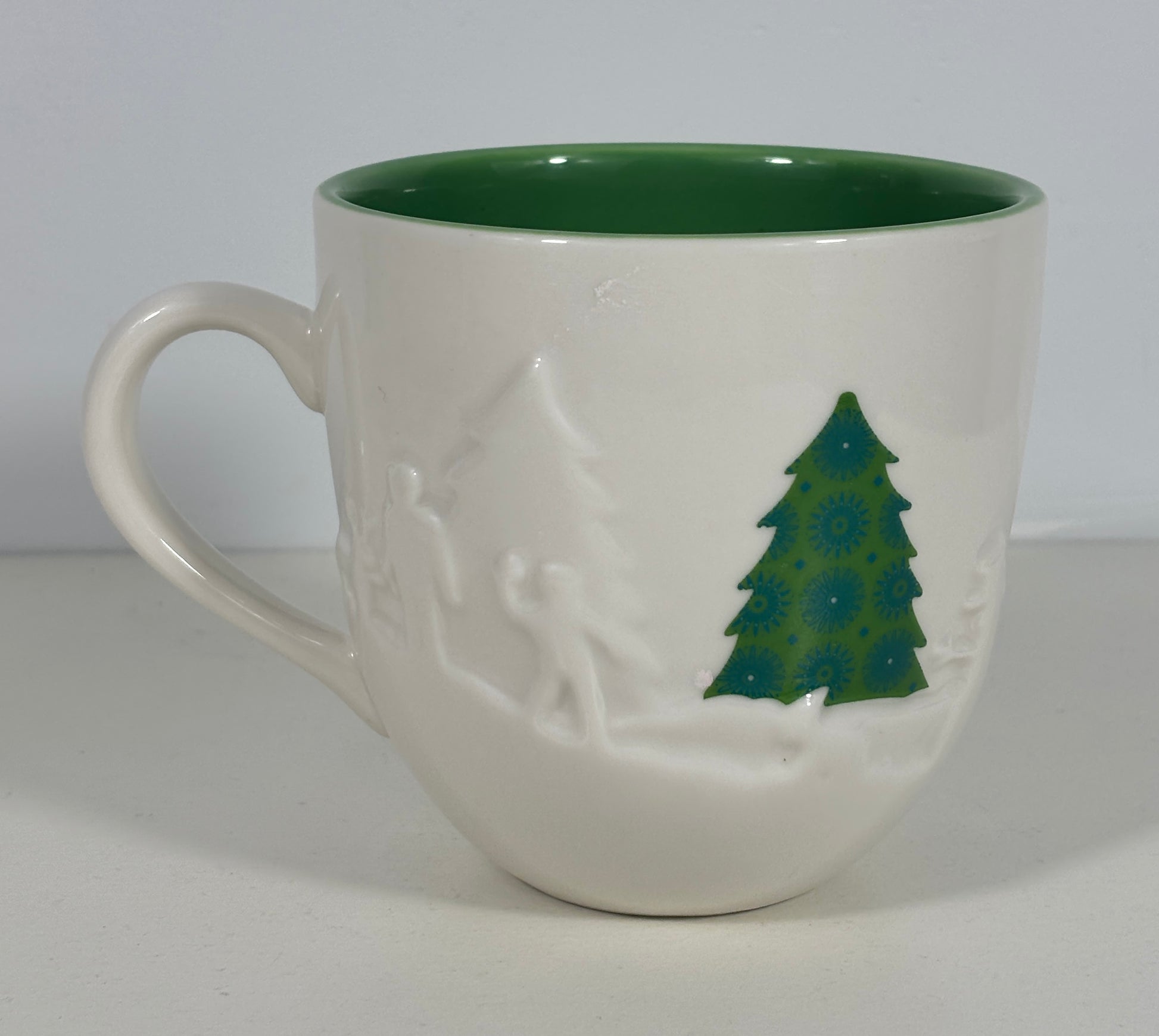A white ceramic mug with a dark green interior and a green Christmas tree design on the side, set against a plain light gray background.