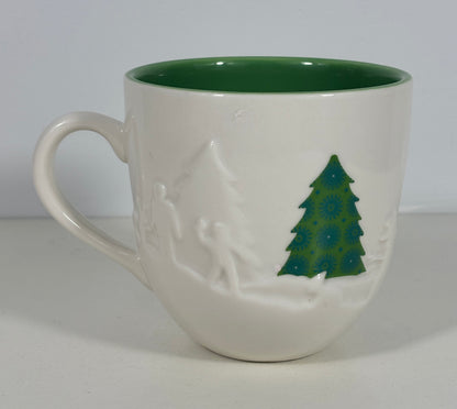 A white ceramic mug with a dark green interior and a green Christmas tree design on the side, set against a plain light gray background.