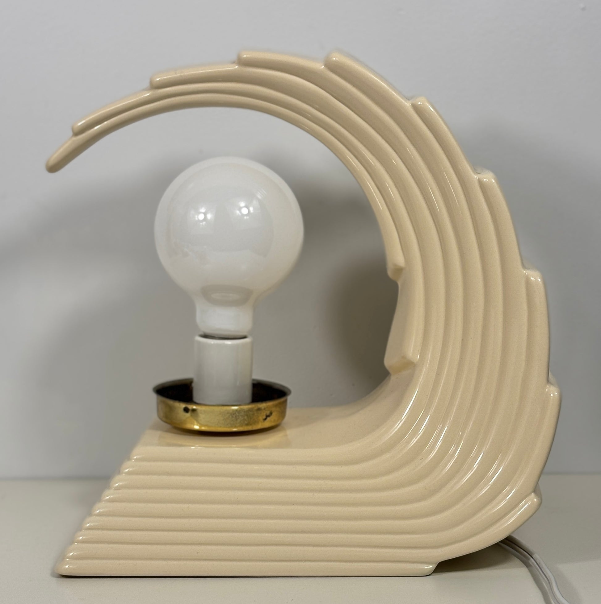 A retro-style table lamp with a cream-colored ceramic wave-shaped base and a matching curved backplate holding a white globe bulb with a gold-toned collar, set against a plain light gray background.