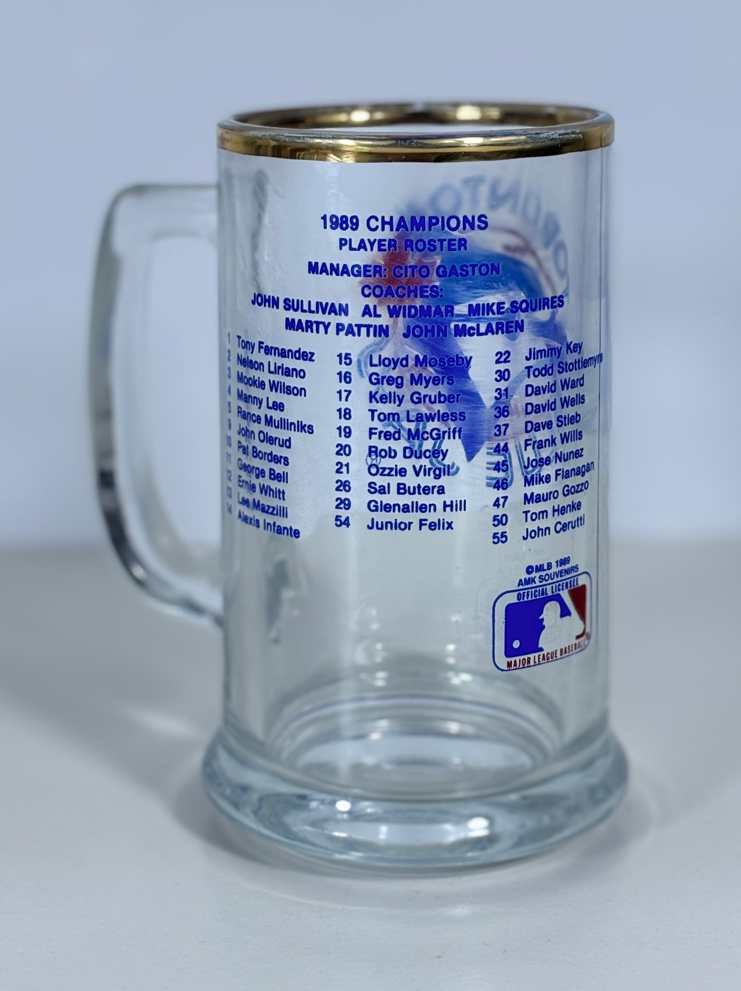 A clear glass beer stein with a gold-colored rim, featuring printed blue text listing the 1989 MLB Champions player roster and a MLB logo near the bottom, photographed against a light gray background.