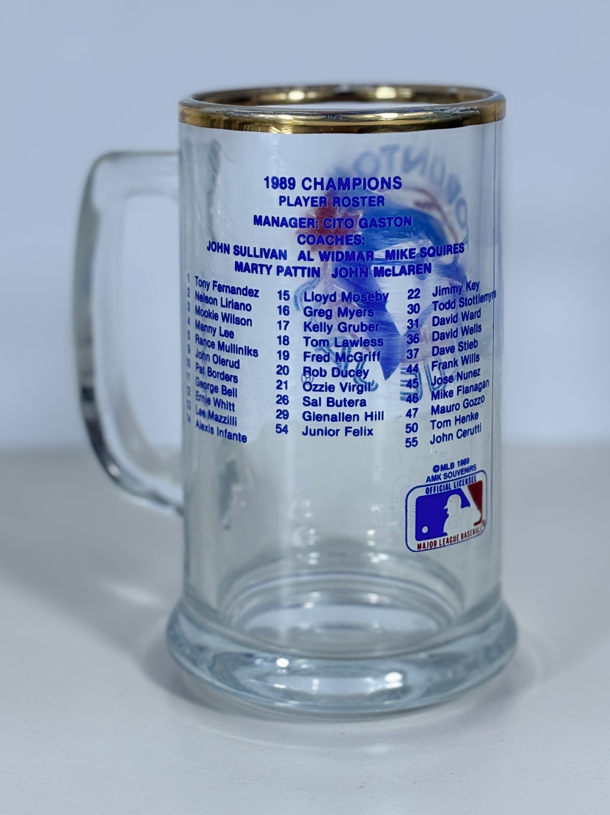 A clear glass beer stein with a gold-colored rim, featuring printed blue text listing the 1989 MLB Champions player roster and a MLB logo near the bottom, photographed against a light gray background.