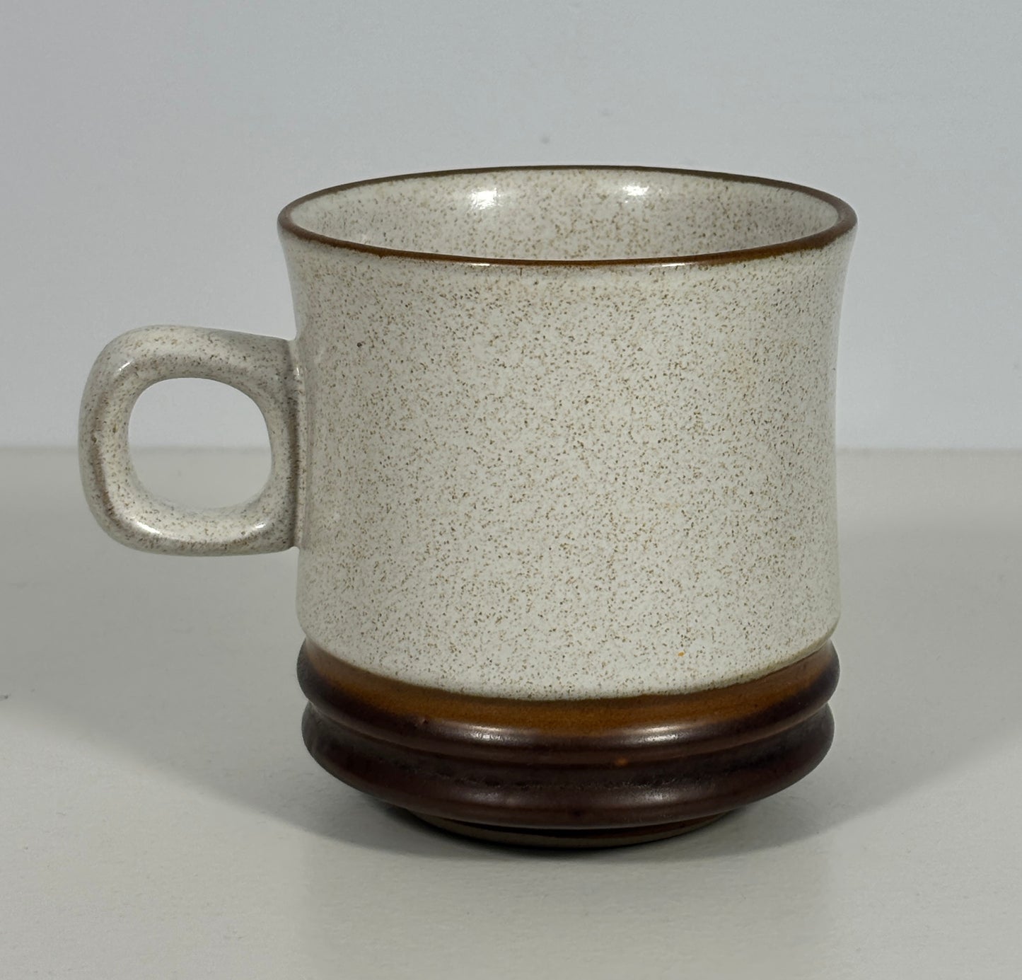 A speckled ceramic mug with a textured off-white body, a dark brown rim, a matching handle, and a glossy dark brown base, shown on a neutral light gray background.