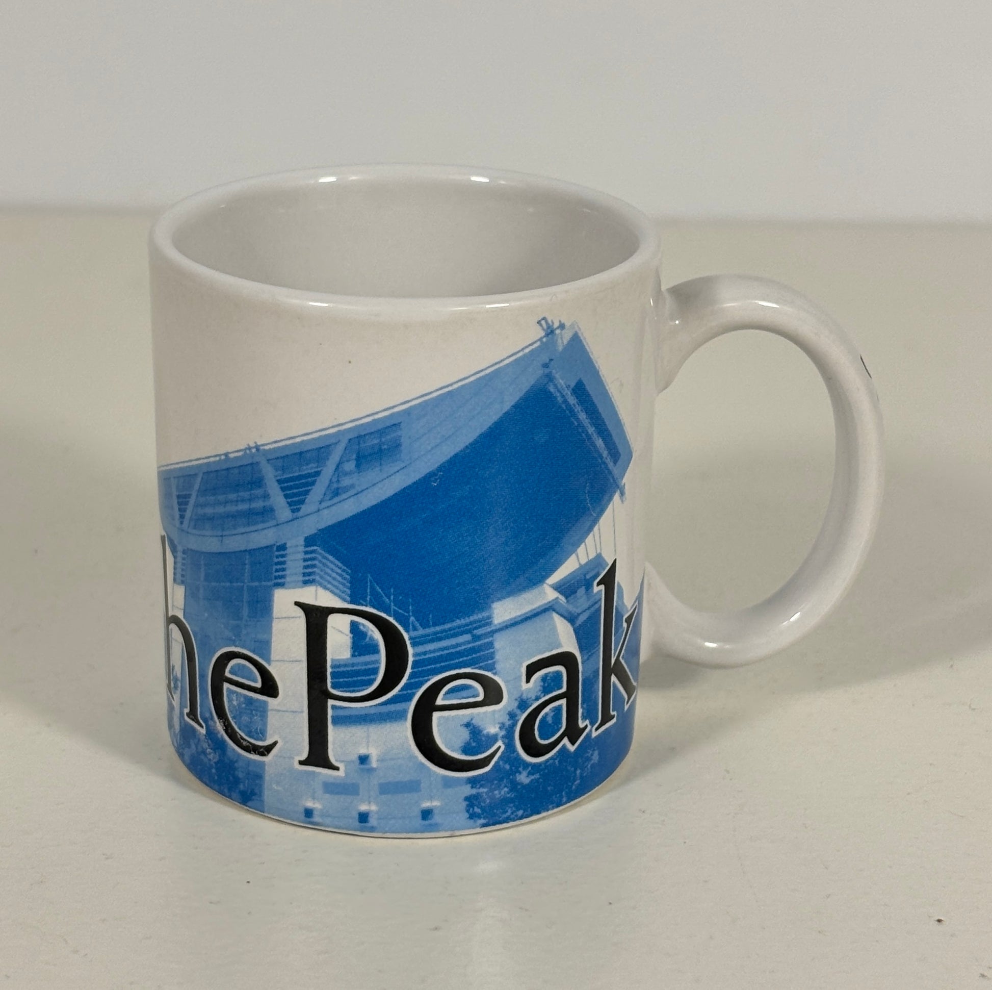 A white ceramic coffee mug featuring a blue graphic of a stadium and the partial text 'The Peak' in black, set against a plain light background.