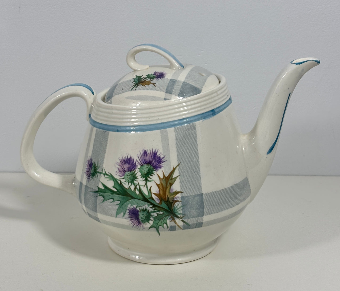 A vintage ceramic teapot with a white body, blue stripes, and a purple thistle floral design, shown on a plain light gray background.