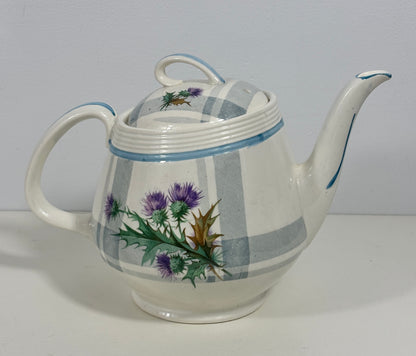 A vintage ceramic teapot with a white body, blue stripes, and a purple thistle floral design, shown on a plain light gray background.