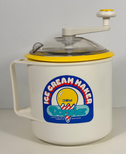 A vintage manual ice cream maker with a white plastic container, yellow lid rim, clear plastic lid, and a hand crank on top. The front features a colorful 'Gratinol Davyier' logo.