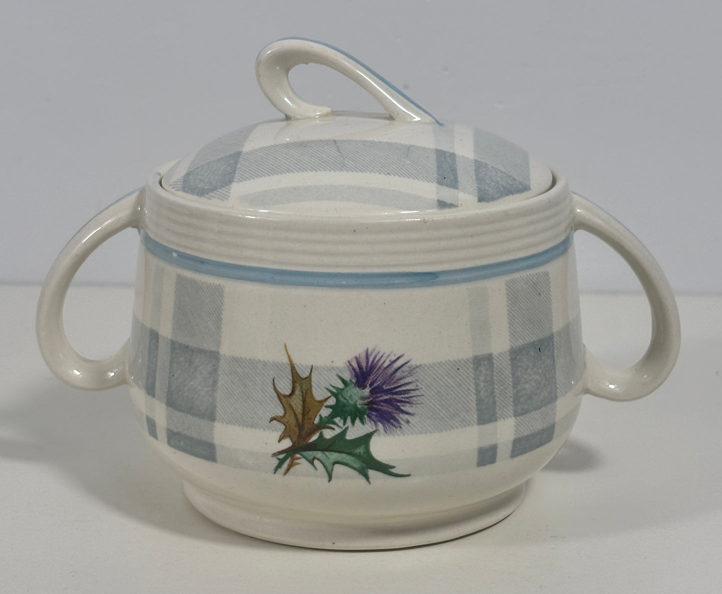 A ceramic lidded sugar bowl with two side handles, featuring a blue and white plaid pattern and a painted thistle and holly design on the front, set against a plain light gray background.