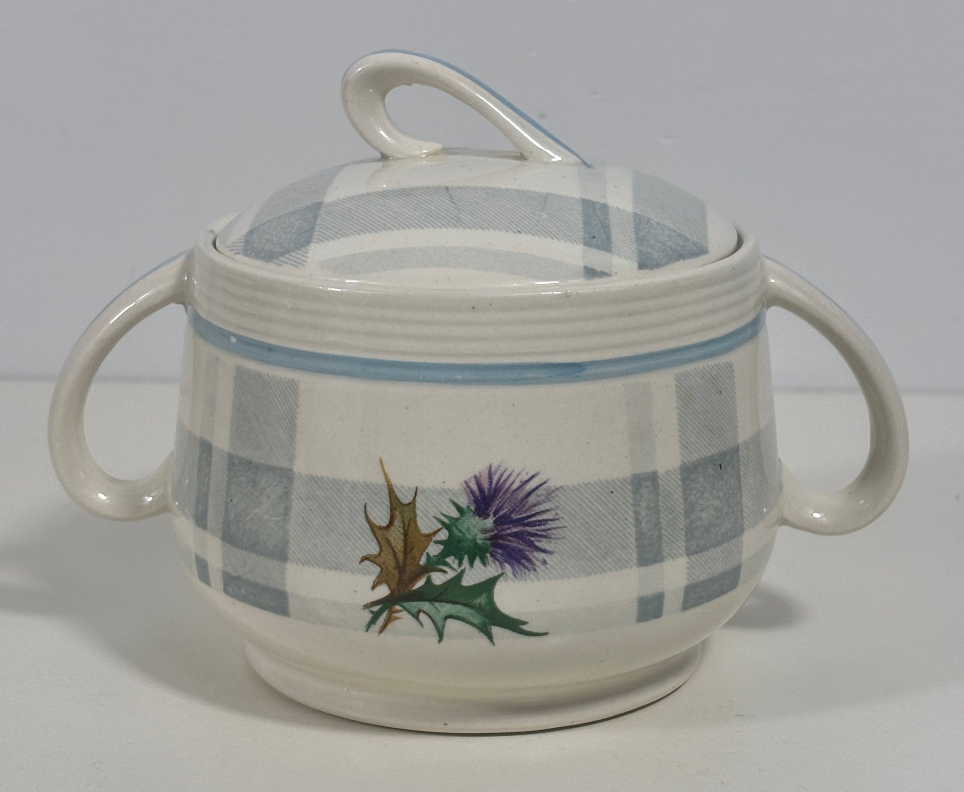 A ceramic lidded sugar bowl with two side handles, featuring a blue and white plaid pattern and a painted thistle and holly design on the front, set against a plain light gray background.