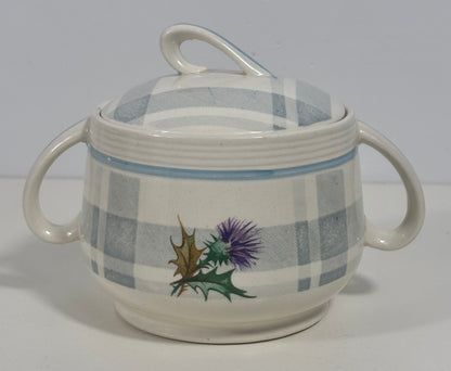 A ceramic lidded sugar bowl with two side handles, featuring a blue and white plaid pattern and a painted thistle and holly design on the front, set against a plain light gray background.