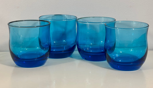 Four translucent cobalt blue glass tumblers with a rounded, wavy shape, arranged on a white surface against a light gray background.