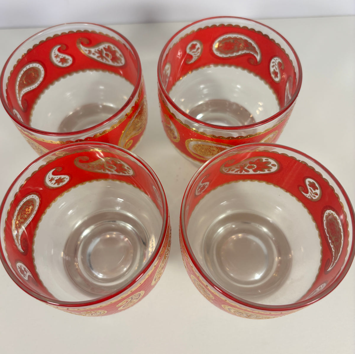 Four clear glass tumblers with red and gold paisley patterns, arranged in a square on a white surface.
