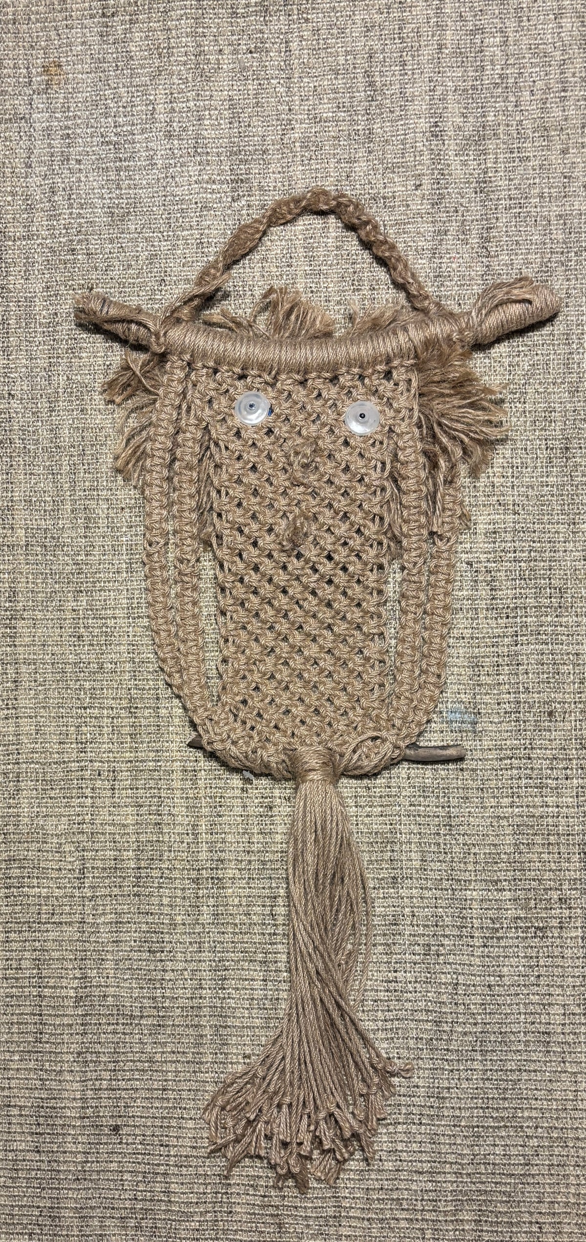 A handmade macrame owl wall hanging made from natural tan jute rope, featuring embroidered white eyes and a hanging loop at the top, displayed against a textured beige linen background.