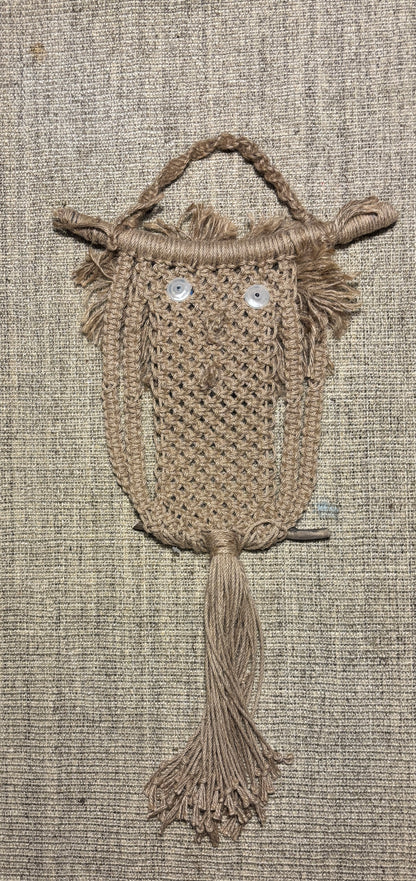 A handmade macrame owl wall hanging made from natural tan jute rope, featuring embroidered white eyes and a hanging loop at the top, displayed against a textured beige linen background.