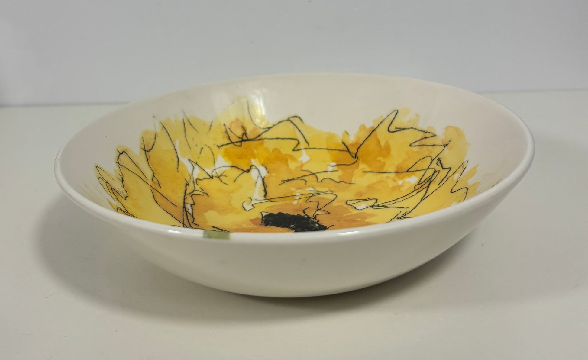 A white ceramic bowl featuring a painted yellow floral design with black and white linework inside, set against a plain light gray background.